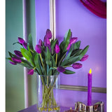 In het Zijden kunstboeket Tulpen in vaas Paars staan levendige paarse tulpen met groene bladeren in een vaas van helder glas, uitgestald op een paars vlak naast een brandende kaars en een decoratieve bak, tegen een lavendelblauwe achtergrond met metallic accenten.