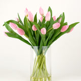 Het Zijden kunstboeket Tulpen in vaas Roze heeft roze tulpen met lange stelen en groene bladeren in een heldere glazen vaas, tegen een effen witte achtergrond.