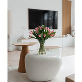 Een moderne woonkamer met een wit vloerkleed, een grote ovale salontafel en een kleine ronde houten bijzettafel. Op de tafel staat het Zijden kunstboeket Tulpen in vaas Roze; een tv en een minimalistisch decor maken de achtergrond compleet.