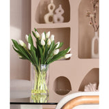 Het Zijden kunstboeket Tulpen in vaas Wit, met witte tulpen en groene bladeren, staat op een glazen tafel. Daarachter staan op beige planken moderne keramische decorstukken en een bloeiende tak in een witte vaas.