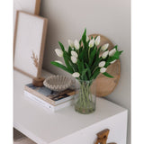 Het Zijden kunstboeket Tulpen in vaas Wit staat op een witte tafel naast gestapelde boeken, een schelpvormig decorstuk en ingelijste kunst tegen een lichtgekleurde muur.