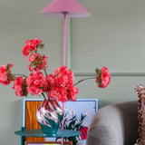 Een roze lamp, een gestreepte vaas met het Zijden kunstbloemenboeket Kersenbloesem Koraal, kleurrijke kunstwerken en een grijze stoel staan tegen een lichtgroene muur, waardoor een gezellige, levendige hoek ontstaat.