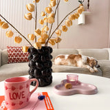 Een gezellige woonkamer met een beige bank, een slapende kat, een sculpturale zwarte vaas met daarin de Kunstbloem Physalis Lampion 90 cm bruin, een roze "LOVE" mok, een boek, een kaars en een koekje op de salontafel.
