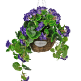 De Kunstbegonia paars in mand UV 70cm is een hangende rieten mand met groene bladeren en levendige paarse bloemen, opgehangen aan een zilveren ketting en voorzien van een klein metalen label met de tekst "Flowers.