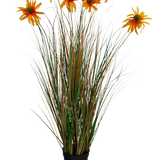De Kunstgras chrysant uienpot 90cm brandvertragend heeft hoge, groene en bruine grasachtige bladeren met zeven oranje kunstmadeliefbloemen in een zwarte pot, tegen een witte achtergrond.