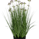 De Kunstgras Prei Bloem Ui Graspot 60cm brandvertragend heeft hoge, dunne groene grasachtige bladeren en delicate kleine paarse bloemen in een zwarte pot, tegen een witte achtergrond.