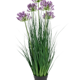 De Kunstgras paarse Agapanthus uiengraspot is een 90 cm hoge, brandvertragende kunstplant met lange groene bladeren en trossen paarse bloemen, weergegeven in een effen zwarte pot tegen een witte achtergrond.