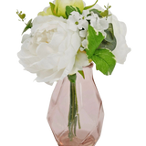 Het Kunstboeket in glas 25 cm heeft een klein boeket van witte bloemen met groene bladeren, gerangschikt in een facetgeslepen, lichtroze glazen vaas.