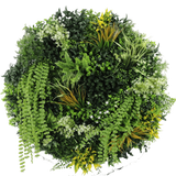 De Kunsthaag op Zwart Frame Ø100 cm heeft een cirkelvormig arrangement van verschillende groene planten en gebladerte, waaronder varens, slingerende wijnranken en bladeren met gele punten, met verschillende bladvormen en texturen tegen een witte achtergrond.