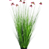 De Kunst Grasplant Chrysant 150 cm brandvertragend heeft lange, dunne groene grasachtige bladeren en lange stelen met roze margrietachtige bloemen, gepresenteerd in een pot tegen een effen witte achtergrond.